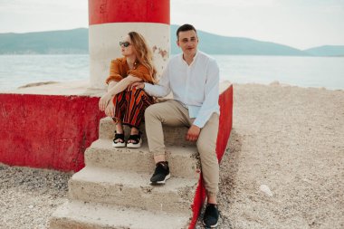 Sahildeki deniz fenerinin yanında, denizin ve dağların arkasında bir çift oturuyor. Tatil aşkı, balayı, seyahat. şık bir çift.