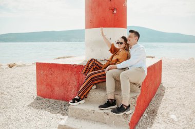 Bir çift, deniz fenerinin yanında denizin ve dağların arka planına karşı oturuyor. Tatil aşkı, balayı, seyahat. Tarz sahibi çift gülüyor ve el sallıyor.