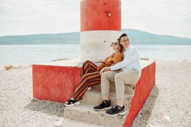 Sahildeki deniz fenerinin yanında, denizin ve dağların arkasında bir çift oturuyor. Tatil aşkı, balayı, seyahat. şık bir çift.