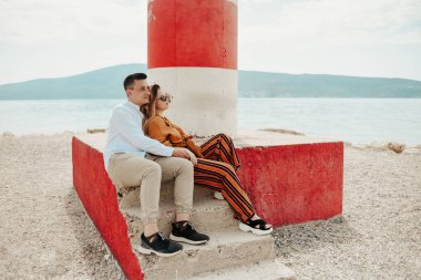 Sahildeki deniz fenerinin yanında, denizin ve dağların arkasında bir çift oturuyor. Tatil aşkı, balayı, seyahat. şık bir çift.