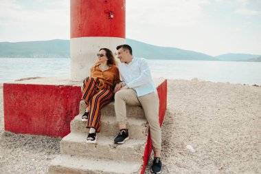 Sahildeki deniz fenerinin yanında, denizin ve dağların arkasında bir çift oturuyor. Tatil aşkı, balayı, seyahat. Tarz sahibi çift gülüyor ve konuşuyor..