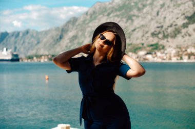 Güneş gözlüklü ve şapkalı bir kadın denizin ve dağların arkasında duruyor, gülümsüyor. Tatil ve açık hava eğlencesi. Tatilde uzun saçlı bir kız. Moda.