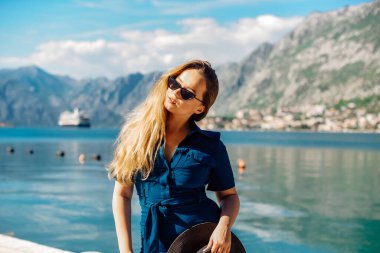 Güneş gözlüklü ve şapkalı bir kadın denizin ve dağların arkasında duruyor. Tatil ve açık hava eğlencesi. Uzun saçlı kız tatilde.
