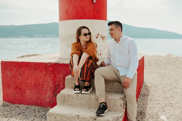 一对夫妇坐在海滩上一座灯塔旁边 面对着大海和群山 度假爱 度蜜月 时髦的夫妇 — 图库照片