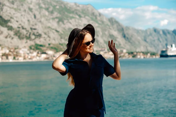 A woman in sunglasses and a hat stands against the backdrop of the sea and mountains, smiling. Vacation and outdoor recreation. Girl with long hair on vacation. Fashion