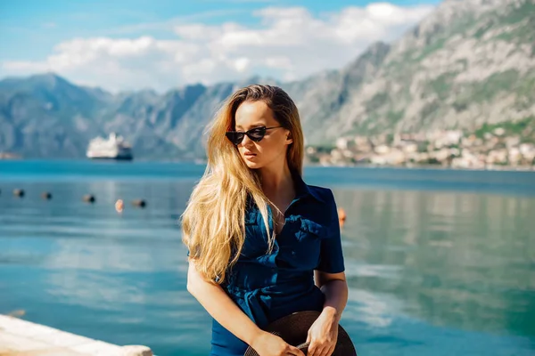 A woman in sun glasses and a hat stands against the backdrop of the sea and mountains. Vacation and outdoor recreation. Girl with long hair on vacation