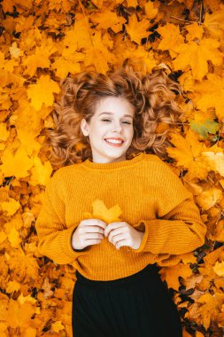 A beautiful girl lies in autumn leaves in the park, gorgeous hair, a teenager smiles and looks at the camera in autumn