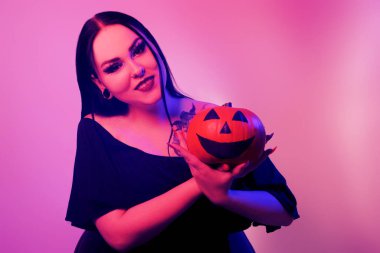 A beautiful brunette holds a pumpkin in her hands and looks at it. Woman with Halloween makeup, dark lipstick. Model in neon pink
