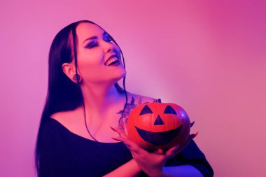 A beautiful brunette holds a pumpkin in her hands and looks at it. Woman with Halloween makeup, dark lipstick. Model in neon pink