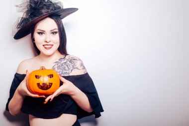 Bir cadı kız elinde bir balkabağı tutuyor ve kameraya doğru uzanıyor, bir kadın Cadılar Bayramı 'nı kutluyor, beyaz bir arka planda fotoğrafı var.