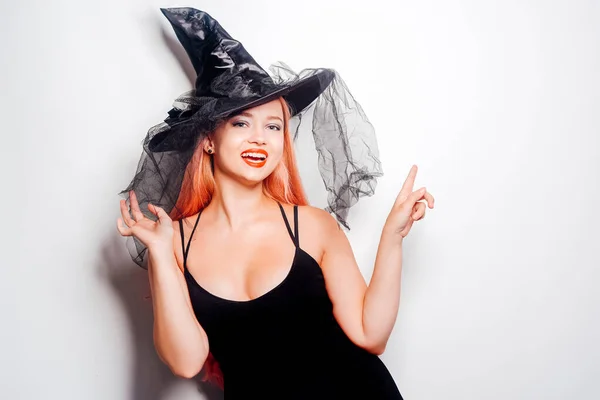 Pink-haired woman witch, in a hat on a white background, halloween costume. The girl smiles and looks to the side at the empty space. Halloween party