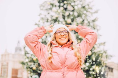 Noel ağacının arka planında gözlüklü bir kızın şirin portresi. Kış, ilk kar, pembe ceketli bir sarışın ve beyaz şapkalı bir gülümseme. Yeni Yıl.