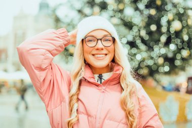 Noel ağacının arka planında gözlüklü bir kızın şirin portresi. Kış, ilk kar, pembe ceketli bir sarışın ve beyaz şapkalı bir gülümseme. Yeni Yıl.