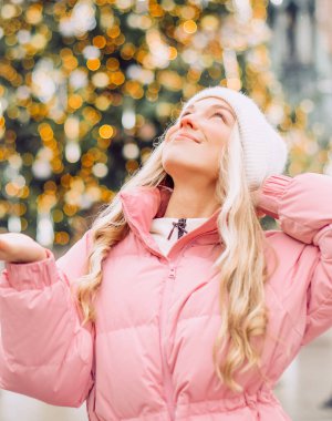Sokakta bir Noel ağacının arka planında duran şapkalı ve pembe ceketli hoş bir sarışın. Kış yürüyüşü, yeni yıl, bayram günü