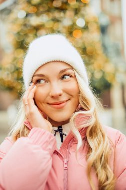Sokakta bir Noel ağacının arka planında duran şapkalı ve pembe ceketli hoş bir sarışın. Kış yürüyüşü, yeni yıl, bayram günü.