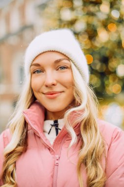 Sokakta bir Noel ağacının arka planında duran şapkalı ve pembe ceketli hoş bir sarışın. Kış yürüyüşü, yeni yıl, bayram günü.