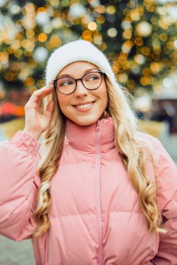 Noel ağacının arka planında gözlüklü bir kızın şirin portresi. Kış, ilk kar, pembe ceketli bir sarışın ve beyaz şapkalı bir gülümseme. Yeni Yıl.