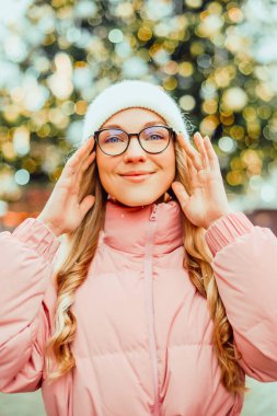 Noel ağacının arka planında gözlüklü bir kızın şirin portresi. Kış, ilk kar, pembe ceketli bir sarışın ve beyaz şapkalı bir gülümseme. Yeni Yıl.
