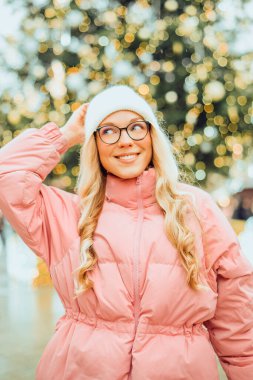 Noel ağacının arka planında gözlüklü bir kızın şirin portresi. Kış, ilk kar, pembe ceketli bir sarışın ve beyaz şapkalı bir gülümseme. Yeni Yıl.