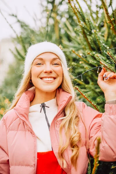 A cute blonde chooses a Christmas tree for the new year. A woman in a hat and pink jacket smiles at the camera in snowy weather. Winter holidays and new year