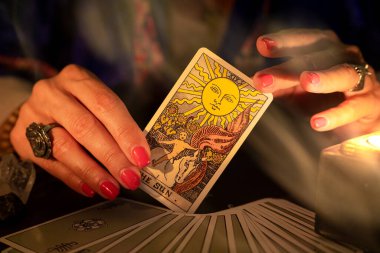 Fortune teller female hands showing The Sun tarot card, symbol of positivity and optimism, during a reading. Close-up with candle light and smoke, moody atmosphere.