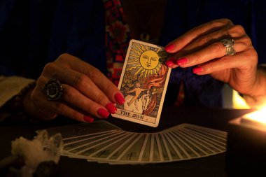 Fortune teller hands showing The Sun tarot card, symbol of positivity, during a reading. Close-up with candle light, moody atmosphere.