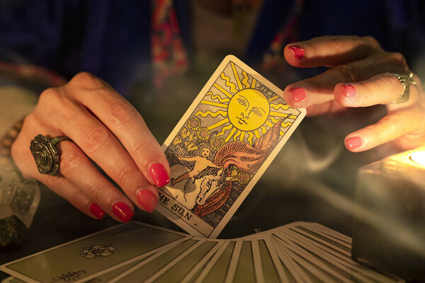 Fortune teller female hands showing The Sun tarot card, symbol of positivity and optimism, during a reading. Close-up with candle light and smoke, moody atmosphere.