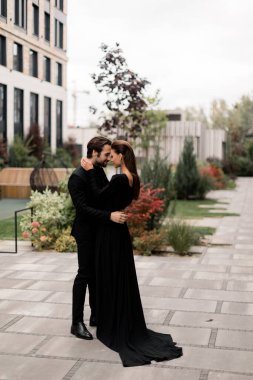 Photoshoot 'ta genç ve moda bir çiftin portresi. Şık takım elbiseli yakışıklı sakallı adam siyah elbiseli muhteşem kadına sarılıyor, fotoğraf için poz veriyor..