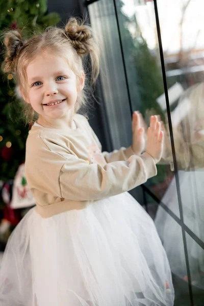 Sarı saçlı, Noel ağacında dikilip kameraya bakan sevimli küçük kız. 