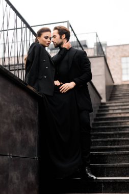 Photoshoot 'ta genç ve moda bir çiftin portresi. Şık takım elbiseli yakışıklı sakallı adam ve şehir merdivenlerinde poz veren siyah elbiseli muhteşem kadın..