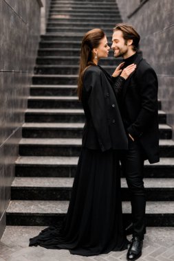 Photoshoot 'ta genç ve moda bir çiftin portresi. Şık takım elbiseli yakışıklı sakallı adam ve şehir merdivenlerinde poz veren siyah elbiseli muhteşem kadın..