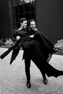 Siyah-beyaz, sokaklarda dans eden, sarılan, şık siyah görünümlü mutlu evli çiftin resmi. Yakışıklı sakallı adam muhteşem genç bayana sarılıyor, gülümsüyor, modern düğün kavramı.