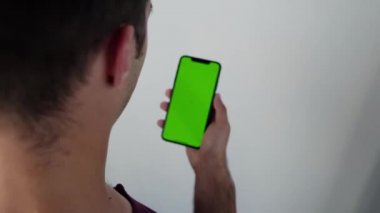 Arkadan çekilen kamera, elinde cep telefonu olan, elinde yeşil ekran, arkasında Krom Anahtar Yeşil Ekran İzleyen Akıllı Telefon olan bir adamın görüntüsünü kaydediyor.