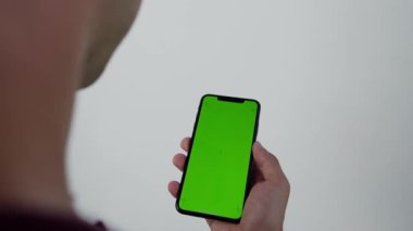 Arkadan çekilen kamera, elinde cep telefonu olan, elinde yeşil ekran, arkasında Krom Anahtar Yeşil Ekran İzleyen Akıllı Telefon olan bir adamın görüntüsünü kaydediyor.