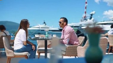 Genç ve mutlu bir çift, bir sahil lokantasında yatlara ve gemilere bakan bir fincan kahve eşliğinde sohbet ederken eğleniyor.