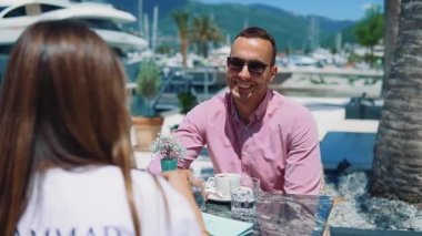 Genç ve mutlu bir çift, bir sahil lokantasında yatlara ve gemilere bakan bir fincan kahve eşliğinde sohbet ederken eğleniyor.