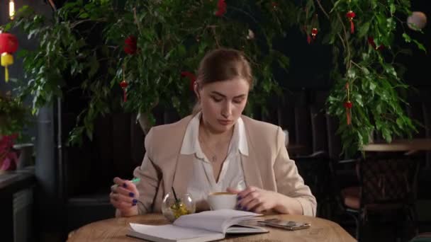 Una chica con el pelo rubio se sienta en una mesa y bebe café con postre — Vídeos de Stock
