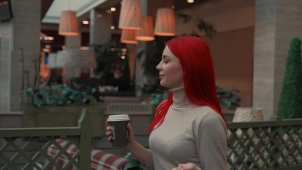Una chica con el pelo rojo camina por el centro comercial con café — Vídeos de Stock