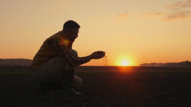 Een man agronomist onderzoekt de grond in zijn handen in een veld bij zonsondergang — Stockvideo