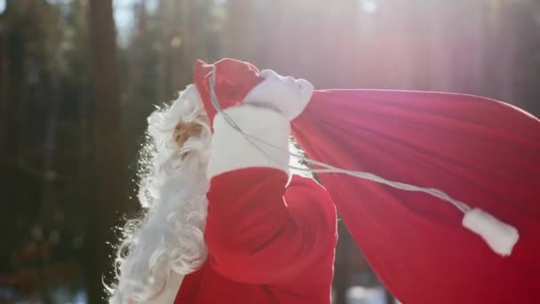 Un retrato de Santa Claus en un parque soleado arroja un saco rojo con regalos en el hombro — Vídeos de Stock