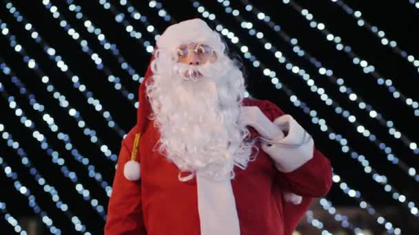 Noel Baba dışarıda yanıp sönen çelenklerin arka planında baş parmağını gösteriyor. — Stok video