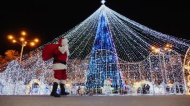 Noel Baba eğlenceli bir şekilde meydandaki Noel ağacının yanında yürüyor.