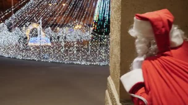 Babbo Natale sbircia dietro l'angolo guardando l'albero di Natale delle città in piazza — Video Stock