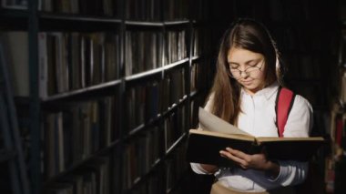 Düşünceli liseli kız kütüphanede duran bir kitap okuyor.