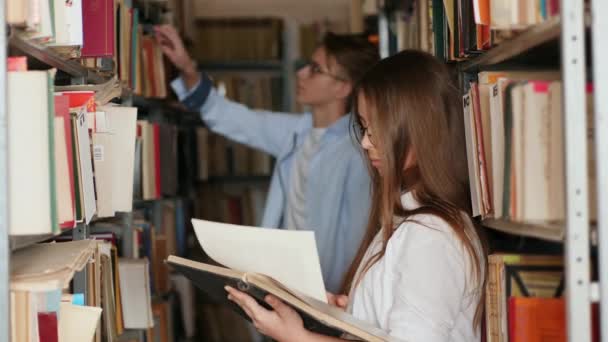 Tinédzserek a városi könyvtárban állnak a könyvespolcok sorai között. — Stock videók