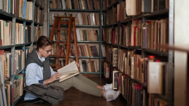 Guy nastolatek czyta książkę w bibliotece siedząc na podłodze między regałami — Wideo stockowe
