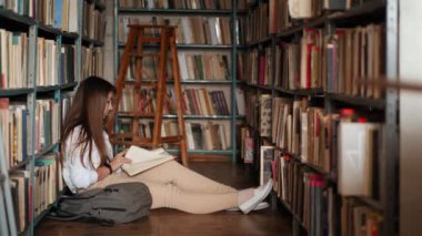 Odaklanmış bir öğrenci kız kütüphanede yerde oturup kitap okuyor.