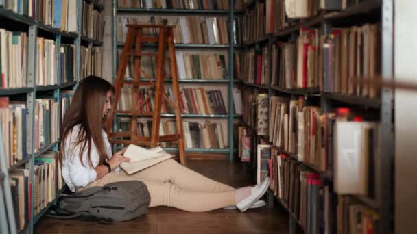 En fokuserad student flicka sitter på golvet och läser bok i biblioteket — Stockvideo