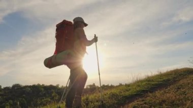 Sırt çantası ve yürüyüş direkleri olan genç bir kadın gün batımında yokuş yukarı tırmanıyor.