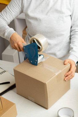 Bir depo çalışanı, yapıştırıcı bant cihazı kullanarak teslimat için bir kutu mal topluyor..